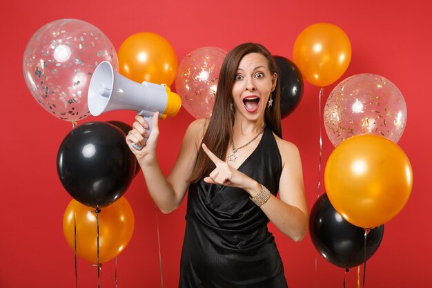 Aufgeregtes junges Mädchen im schwarzen Kleid, das feiert, Megaphon hält und Zeigefinger auf rotem Hintergrund Luftballons beiseite zeigt. Valentinstag, Happy New Year Geburtstag Mockup Urlaub Party Konzept.