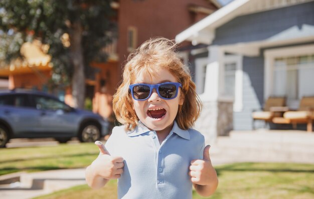 Aufgeregtes Kinderporträt in Sonnenbrille mit Daumen nach oben Schild Nahaufnahme Kopf des niedlichen Kindes im Freien zu Hause nea