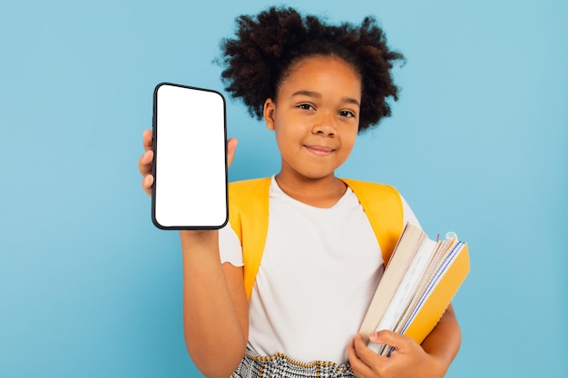 Aufgeregtes kleines afroamerikanisches Schulmädchen, das eine große Zelle in der Hand hält und einen leeren Bildschirm des Telefons auf blauem Studiohintergrund präsentiert, Smartphone-Display-Modell