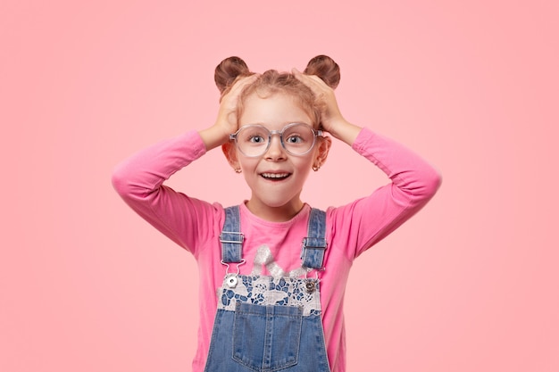 Aufgeregtes kleines Mädchen in lässigem rosa Hemd und Jeansoverall und Brille, die Hände auf dem Kopf hält und mit schockierter Grimasse schaut