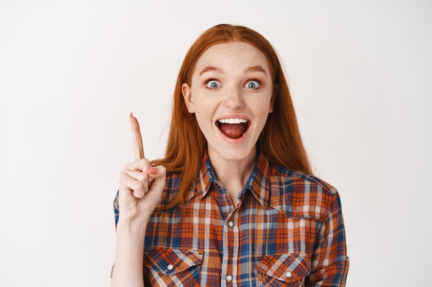 Aufgeregtes rothaariges weibliches Model, das ein ausgezeichnetes Produkt zeigt, mit dem Finger auf die Werbung zeigt, vorne lächelt und über einer weißen Wand steht