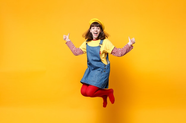 Aufgeregtes Teenager-Mädchen in französischer Baskenmütze, Denim-Sommerkleid, das Daumen hoch springt und herumalbert, isoliert auf gelbem Wandhintergrund im Studio. Menschen Emotionen, Lifestyle-Konzept. Mock-up-Kopienbereich.