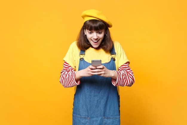 Aufgeregtes Teenager-Mädchen in französischer Baskenmütze, Denim-Sommerkleid mit Handy, SMS-Nachricht isoliert auf gelbem Hintergrund im Studio eingeben. Menschen aufrichtige Emotionen, Lifestyle-Konzept. Mock-up-Kopienbereich.