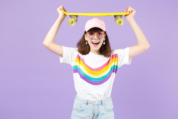 Aufgeregtes Teenager-Mädchen in lebhafter Kleidung, Herzbrille hält gelbes Skateboard über dem Kopf und hält den Mund isoliert auf violettem Hintergrund offen. Menschen aufrichtige Emotionen, Lifestyle-Konzept. Mock-up-Kopienbereich.