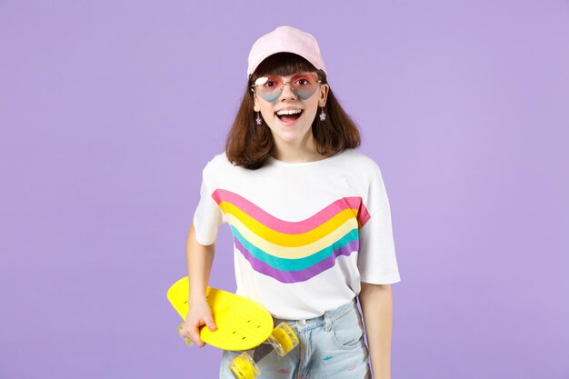 Aufgeregtes Teenager-Mädchen in lebhafter Kleidung, Herzbrille mit gelbem Skateboard, Mund offen haltend isoliert auf violettem Pastellhintergrund. Menschen aufrichtige Emotionen, Lifestyle-Konzept. Mock-up-Kopienbereich.