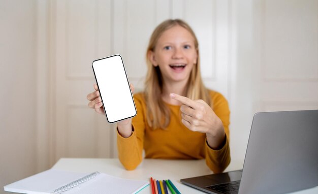 Foto aufgeregtes teenager-mädchen zeigt einen leeren smartphone-bildschirm, während es zu hause studiert