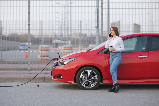 Aufladen des Elektroautos in der Straße. Ökologisches Auto angeschlossen und Batterien aufgeladen. Mädchen verwenden Kaffeegetränk, während sie SmartPhone verwenden und auf die Stromversorgung warten. Zum Aufladen mit Elektrofahrzeugen verbinden