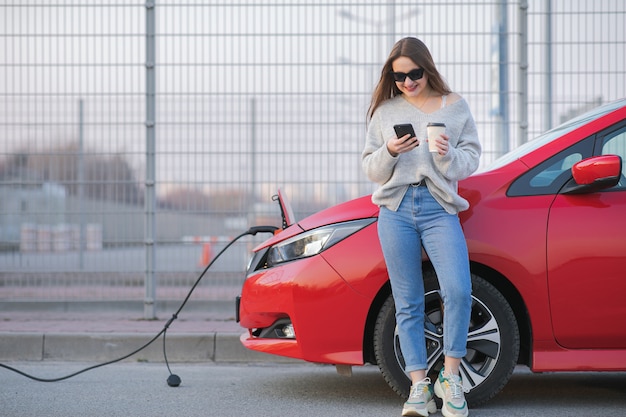 Aufladen des Elektroautos in der Straße. Ökologisches Auto angeschlossen und Batterien aufgeladen. Mädchen verwenden Kaffeegetränk, während sie SmartPhone verwenden und auf die Stromversorgung warten. Zum Aufladen mit Elektrofahrzeugen verbinden