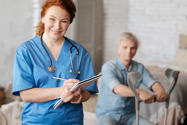 Foto aufmerksamer zuhörer. fähige freundliche medizinische mitarbeiterin, die ihren patienten zu hause besucht und seine symptome aufschreibt, um ihn richtig zu diagnostizieren