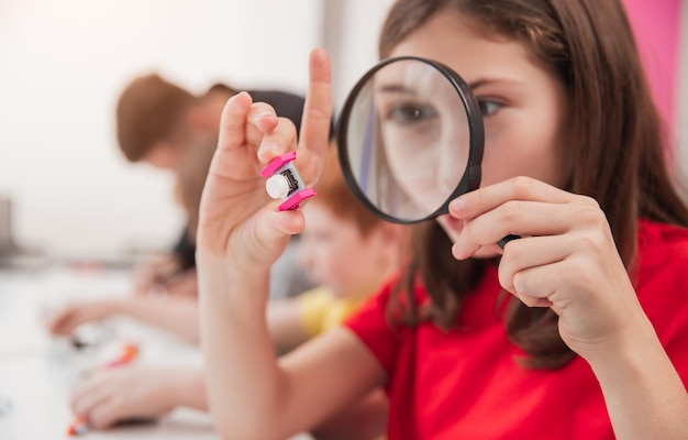 Aufmerksames intelligentes Schulmädchen, das elektronische Details durch eine Lupe betrachtet, während sie Robotik während des Unterrichts in der modernen Schule studiert