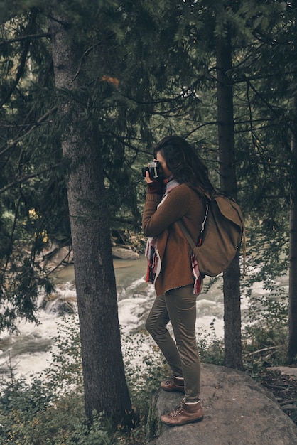 Aufnahme der Ansicht. Volle Länge der jungen modernen Frau mit Rucksack, die die Natur beim Wandern im Wald fotografiert