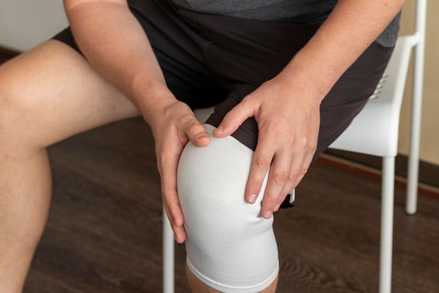 Aufnahme des Mannes, der auf dem weißen Stuhl sitzt und sein Knie mit den Händen hält, elastisches Bandage am rechten Knie Gesundheitswesen