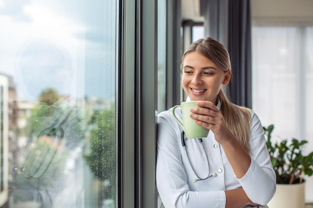 Aufnahme einer Ärztin, die eine Tasse Kaffee hält, während sie sich auf das Fenster freut, das in der Konsultation steht Lächelnde junge Ärztin in weißer medizinischer Uniform und Stethoskop-Blick in die Fernplanung