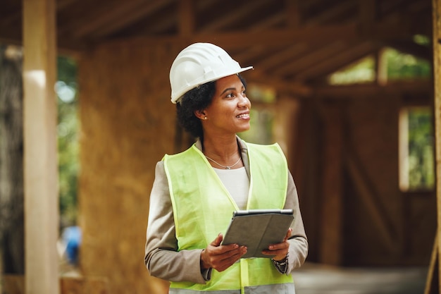 Aufnahme einer afrikanischen Architektin, die ein digitales Tablet benutzt und die Baustelle eines neuen Holzhauses überprüft. Sie trägt Arbeitsschutzkleidung und einen weißen Helm.