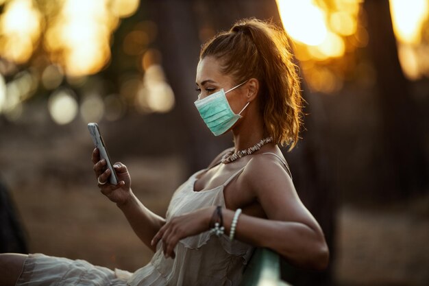 Aufnahme einer attraktiven jungen Frau, die eine chirurgische Maske trägt und ein Smartphone benutzt, während sie sich im Park isoliert.