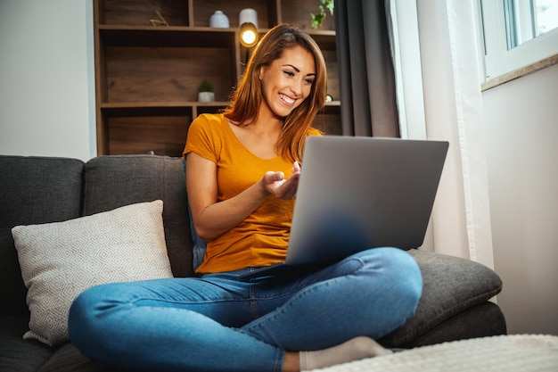 Aufnahme einer attraktiven jungen Frau, die im Schneidersitz auf dem Sofa sitzt und mit ihrem Laptop einen Videoanruf mit jemandem zu Hause führt.