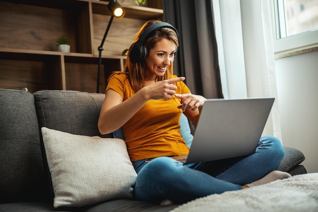 Aufnahme einer attraktiven jungen Frau, die im Schneidersitz auf dem Sofa sitzt und mit ihrem Laptop und ihren Kopfhörern einen Videochat mit jemandem zu Hause führt.