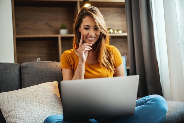 Aufnahme einer attraktiven jungen Frau, die im Schneidersitz auf dem Sofa sitzt und zu Hause ihren Laptop benutzt.