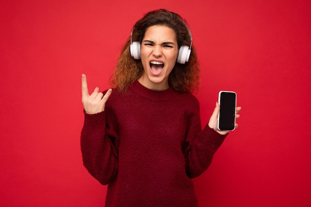 Aufnahme einer attraktiven, positiven, emotionalen jungen, lockigen Frau, die einen dunkelroten Pullover trägt, isoliert auf