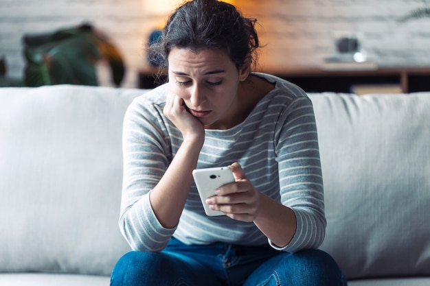 Aufnahme einer besorgten jungen Frau, die ihr Handy benutzt, während sie zu Hause auf dem Sofa im Wohnzimmer sitzt.