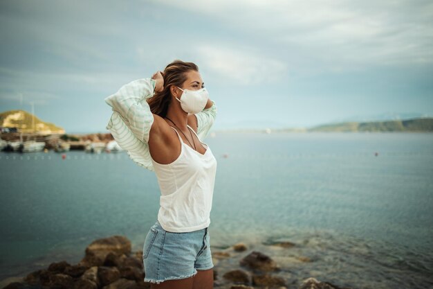 Aufnahme einer entspannten jungen Frau mit N95-Schutzmaske, die während des COVID-19 einen Urlaub am Strand genießt.
