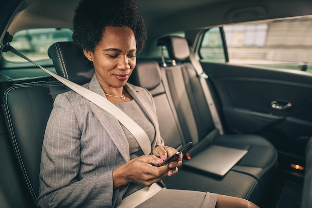 Aufnahme einer erfolgreichen schwarzen Geschäftsfrau, die während ihrer morgendlichen Fahrt auf dem Rücksitz eines Autos ein Smartphone benutzt.