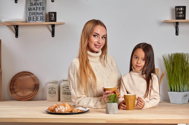 Aufnahme einer fröhlichen Mutter und Tochter, die morgens zusammen am Küchentisch sitzen und heißen Tee trinken.