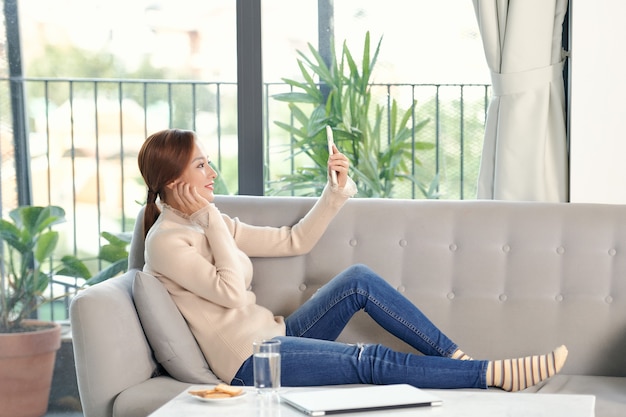 Aufnahme einer glücklichen jungen Asiatin, die ein Selfie mit ihrem Handy macht, während sie im Wohnzimmer sitzt.