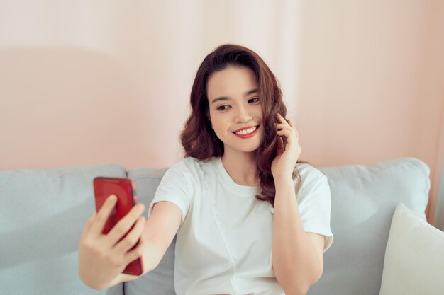 Aufnahme einer glücklichen jungen Asiatin, die mit ihrem Handy ein Selfie macht, während sie im Wohnzimmer sitzt.