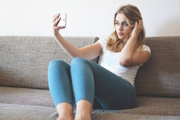 Aufnahme einer glücklichen jungen Frau, die ein Selfie mit ihrem Handy macht, während sie im Wohnzimmer sitzt