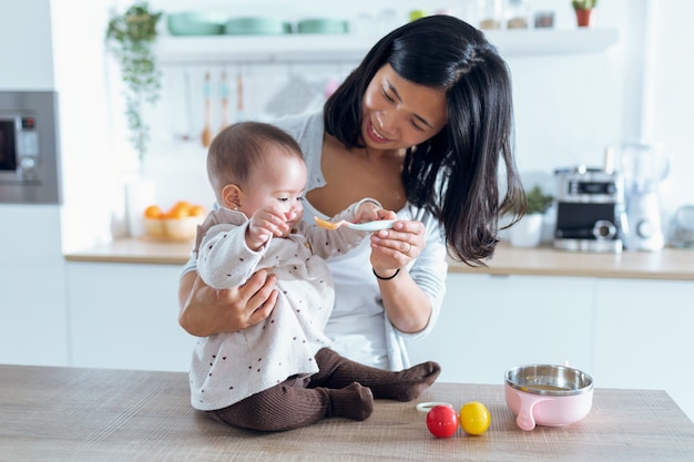 Aufnahme einer glücklichen jungen Mutter, die ihr süßes Baby mit Fruchtbrei in der Küche zu Hause füttert.