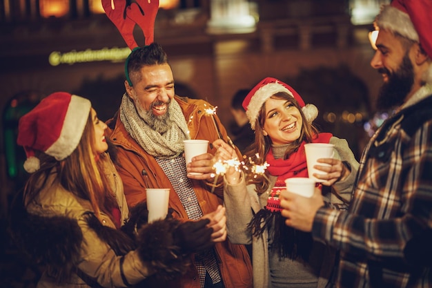 Aufnahme einer Gruppe fröhlicher junger Freunde, die sich mit Wunderkerzen vergnügen und bei einer Abendparty auf dem Weihnachtsmarkt ein heißes Getränk genießen.