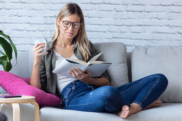 Aufnahme einer hübschen jungen Frau, die ein Buch liest und eine Tasse Kaffee trinkt, während sie zu Hause auf dem Sofa sitzt.