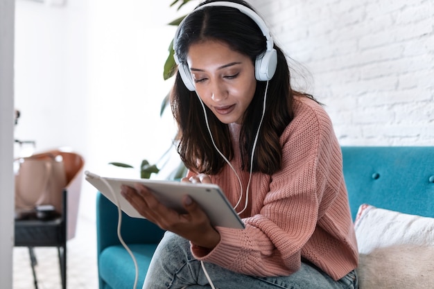 Aufnahme einer hübschen jungen Frau, die mit digitalem Tablet Musik hört, während sie zu Hause auf dem Sofa sitzt.