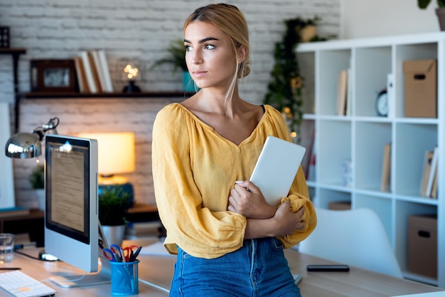 Aufnahme einer intelligenten freiberuflichen Geschäftsfrau, die mit einem digitalen Tablet arbeitet, während sie in ihrem kleinen Startup-Unternehmen durch das Fenster schaut.