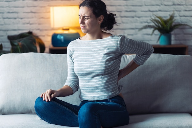 Aufnahme einer jungen Frau mit Rückenschmerzen, die zu Hause auf dem Sofa im Wohnzimmer sitzt.