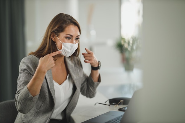 Aufnahme einer jungen Geschäftsfrau mit Schutzmaske, die allein in ihrem Büro sitzt und während einer Corona-Virus-Pandemie einen Videoanruf auf dem Laptop führt.