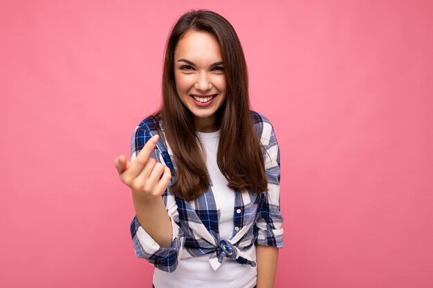 Aufnahme einer jungen, glücklich lächelnden, schönen brünetten Frau mit aufrichtigen Emotionen, die ein trendiges Karohemd einzeln auf rosafarbenem Hintergrund mit leerem Raum trägt.