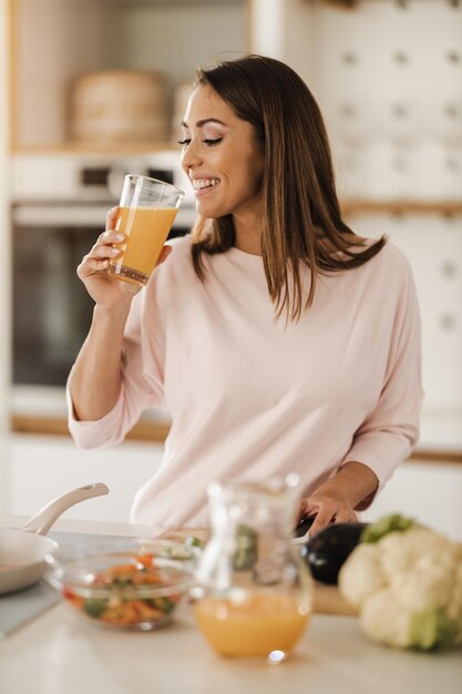 Aufnahme einer jungen lächelnden Frau, die in ihrer Küche frischen Orangensaft trinkt.