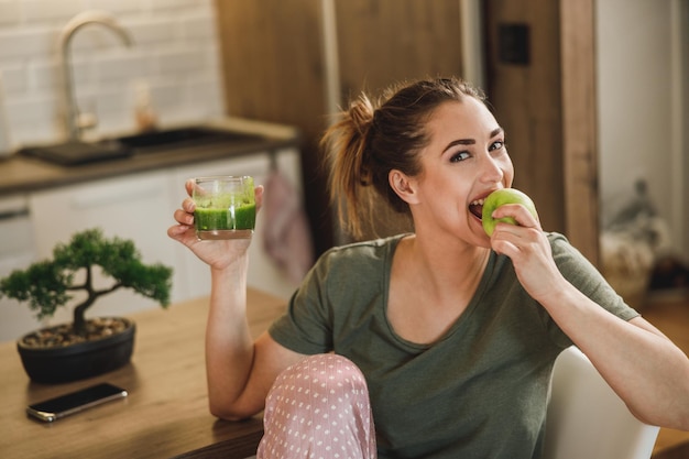 Aufnahme einer jungen lächelnden Frau, die zu Hause Apfelfrüchte isst und grünen Smoothie probiert.