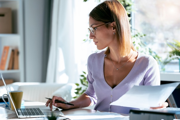 Aufnahme einer konzentrierten jungen Geschäftsfrau, die mit dem Computer arbeitet, während sie zu Hause einige Dokumente im Büro konsultiert.