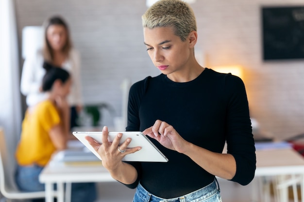 Aufnahme einer konzentrierten jungen Unternehmerin, die ihr digitales Tablet benutzt, während sie im Büro steht.