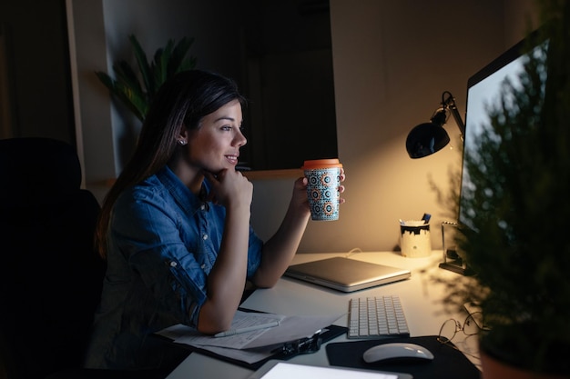 Aufnahme einer lächelnden jungen Frau, die spät in der Nacht Papierkram erledigt und Kaffee trinkt.