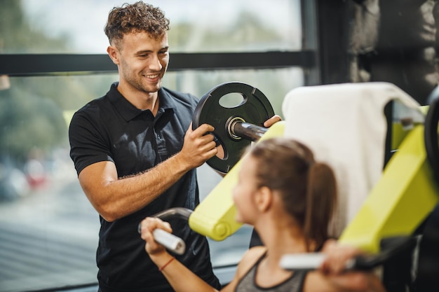 Aufnahme einer muskulösen jungen Frau in Sportkleidung, die mit einem persönlichen Trainer am Fitnessgerät trainiert. Sie pumpt ihre Schultermuskulatur mit schwerem Gewicht auf.