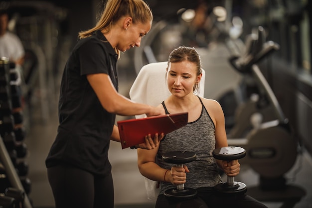 Aufnahme einer muskulösen jungen Frau in Sportkleidung, die mit einer Personal Trainerin im Fitnessstudio trainiert. Sie pumpt ihre Muskeln mit einer Hantel auf.