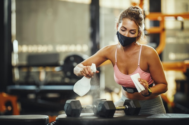 Aufnahme einer muskulösen jungen Frau mit Schutzmaske, die Fitnessgeräte nach dem Training während der Covid-19-Pandemie reinigt.