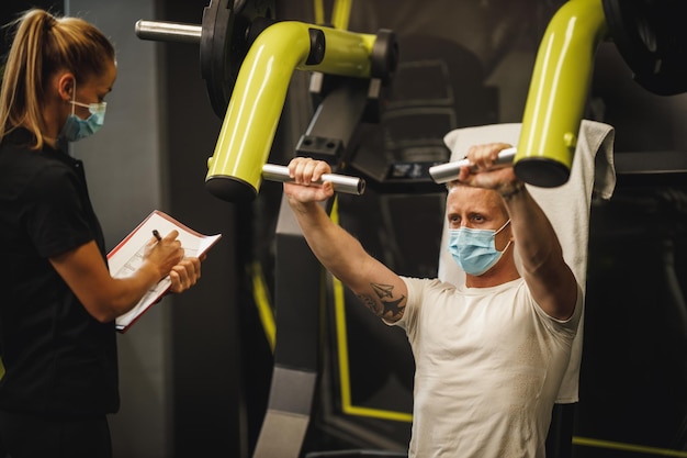Aufnahme einer muskulösen jungen Frau mit Schutzmaske, die während der Covid-19-Pandemie mit einem persönlichen Trainer am Fitnessgerät trainiert. Sie pumpt ihre Brustmuskulatur mit schwerem Gewicht auf.