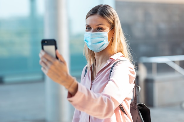 Aufnahme einer schönen blonden Frau mit Gesichtsmaske, die ein Selfie mit dem Smartphone auf der Straße macht.