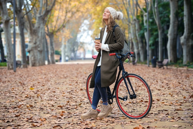 Aufnahme einer schönen Frau mit einem Vintage-Fahrrad, die Kaffee trinkt, während sie im Herbst durch den Park radelt.