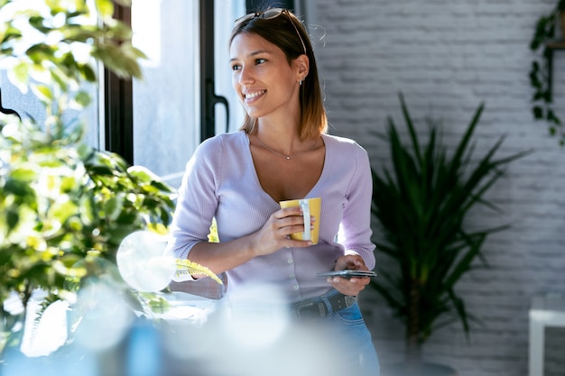 Aufnahme einer schönen jungen Frau, die ihr Handy benutzt, während sie zu Hause eine Tasse Kaffee trinkt, die neben dem Fenster steht.